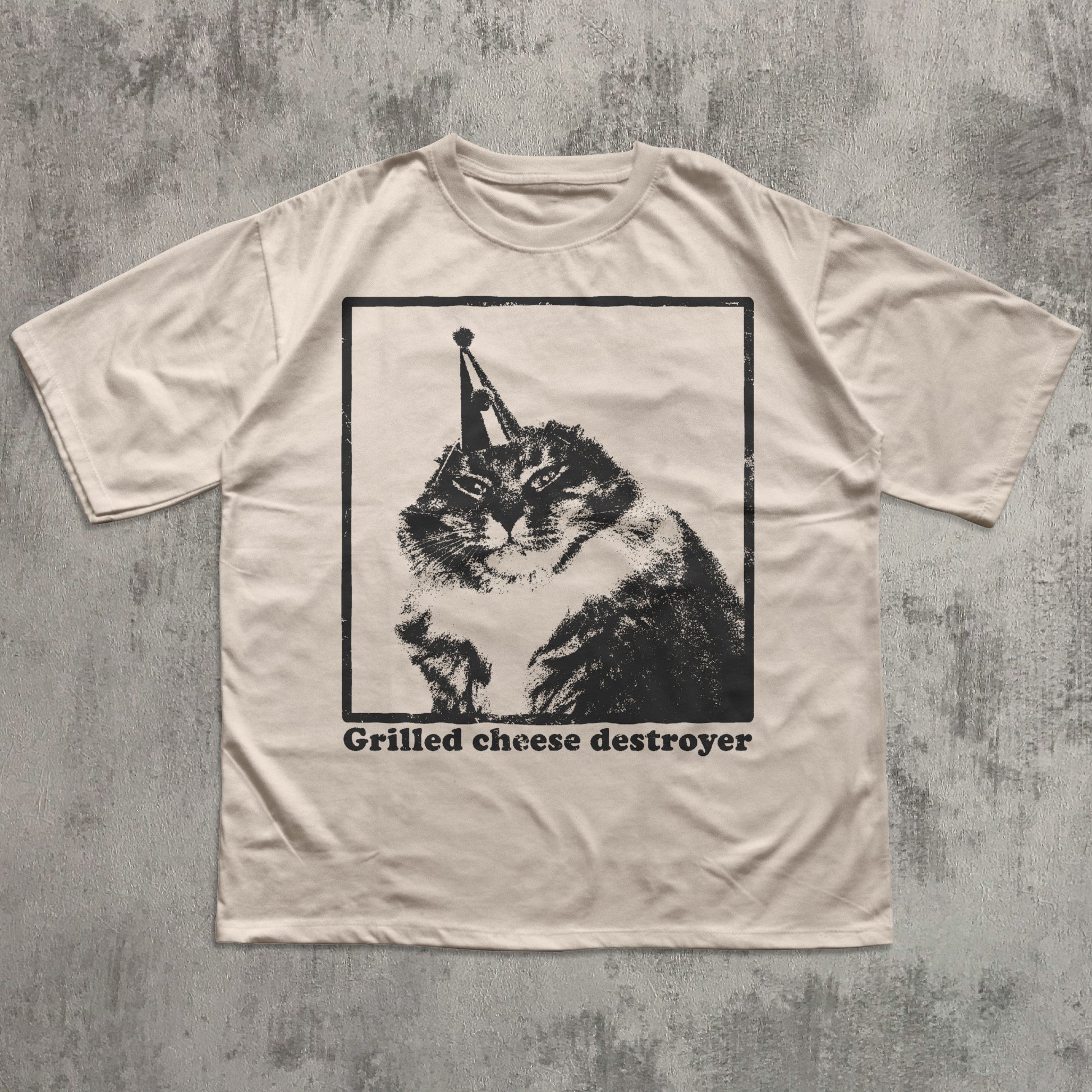 Beige t-shirt with a graphic of a cat wearing a party hat and text 'Grilled cheese destroyer' on a textured gray background.