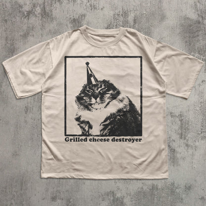 Beige t-shirt with a graphic of a cat wearing a party hat and text 'Grilled cheese destroyer' on a textured gray background.
