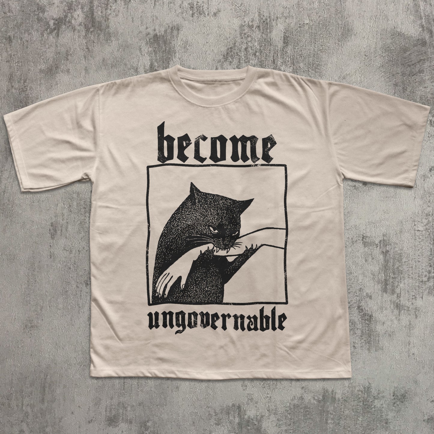 Beige t-shirt with black cat graphic and text 'become ungovernable' on a textured gray background