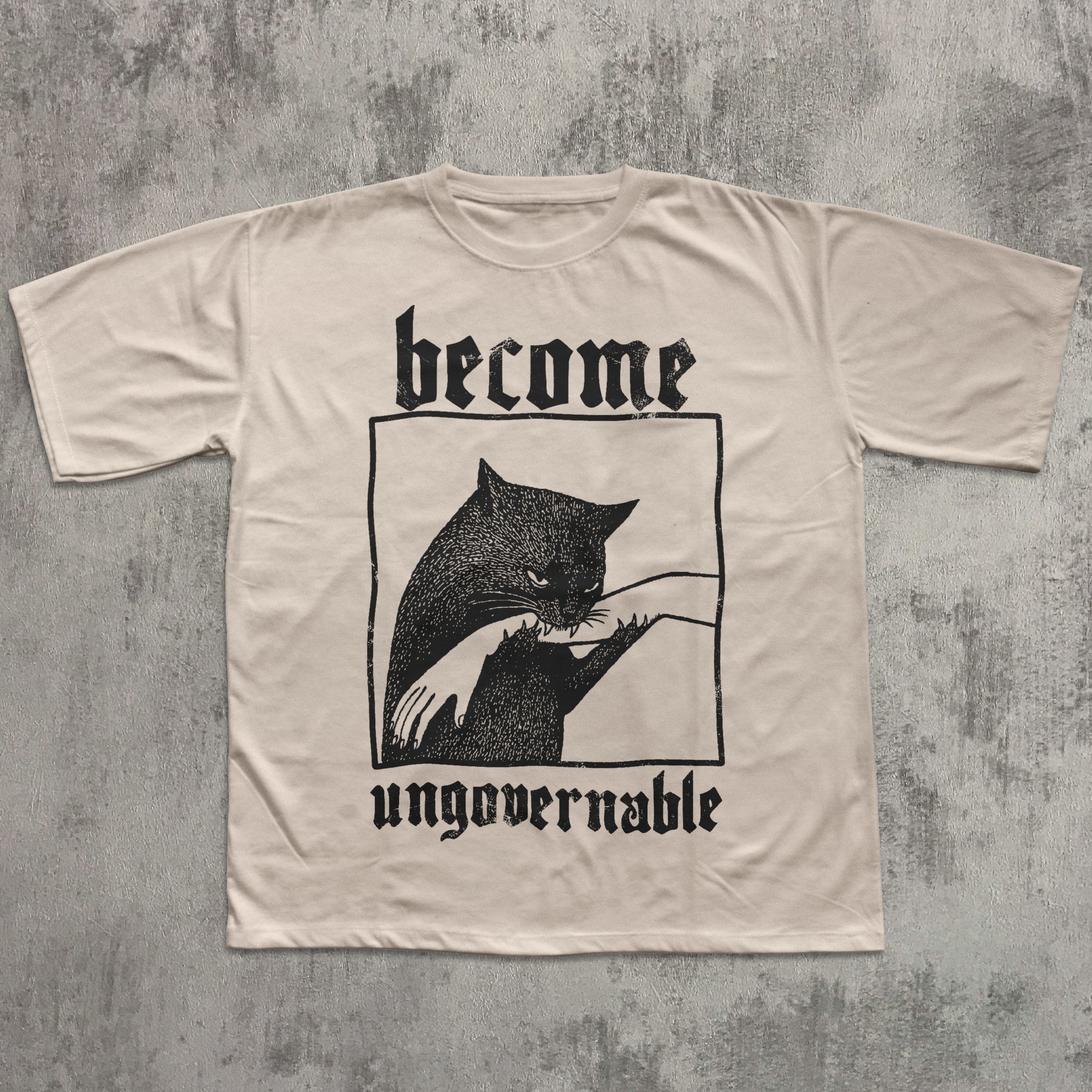 Beige t-shirt with black cat graphic and text 'become ungovernable' on a textured gray background