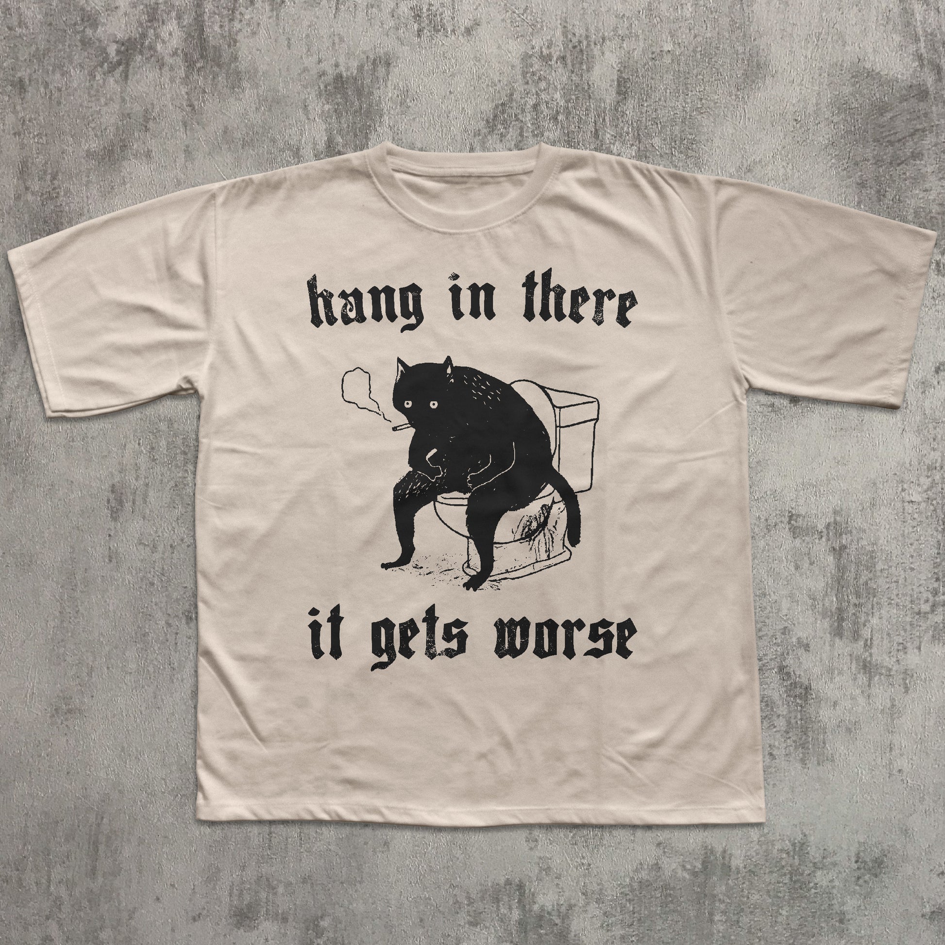 Beige t-shirt with black graphic of a cat and text on a textured gray background