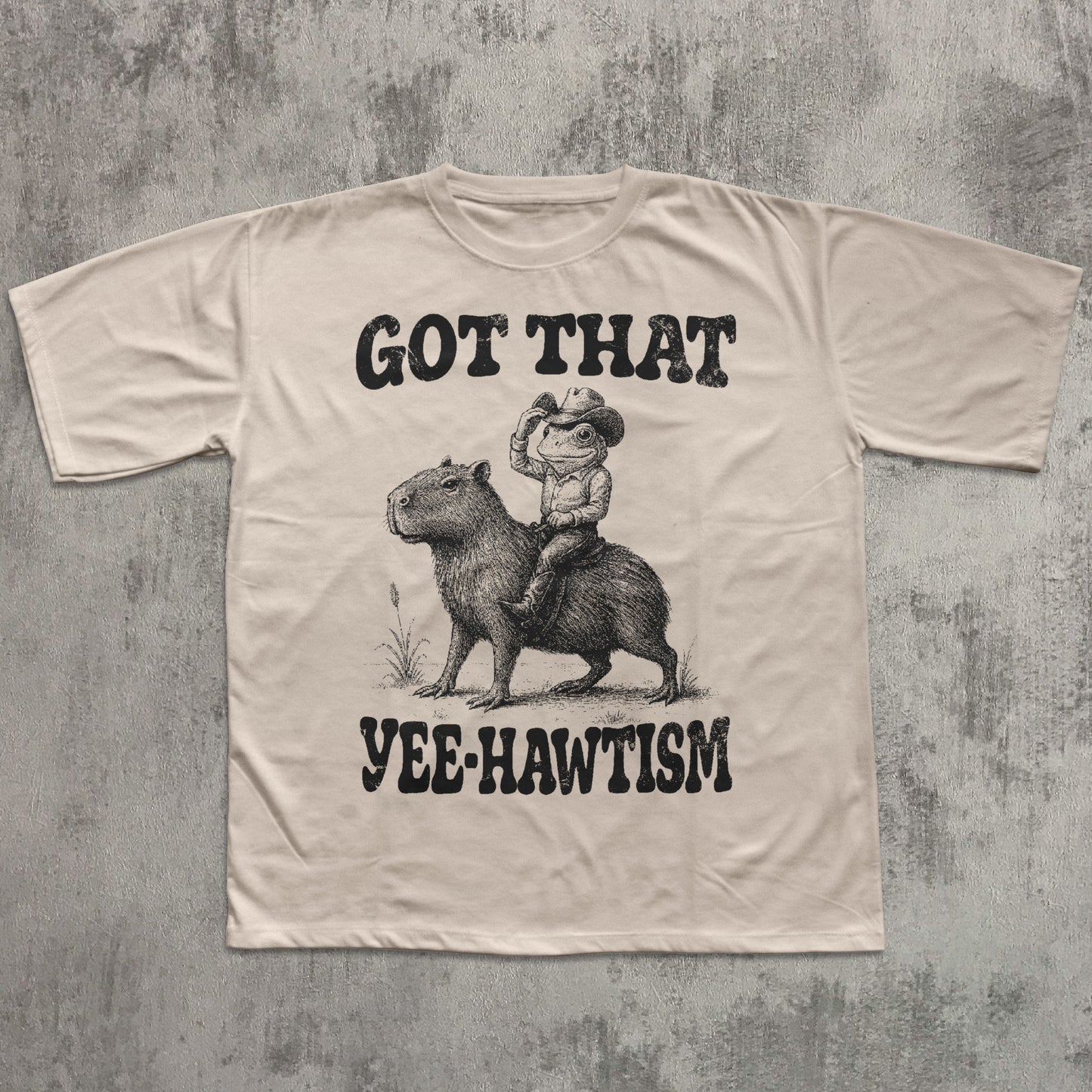 Beige t-shirt with graphic of a cowboy riding a capybara and text 'Got That Yee-Hawtism' on a textured gray background