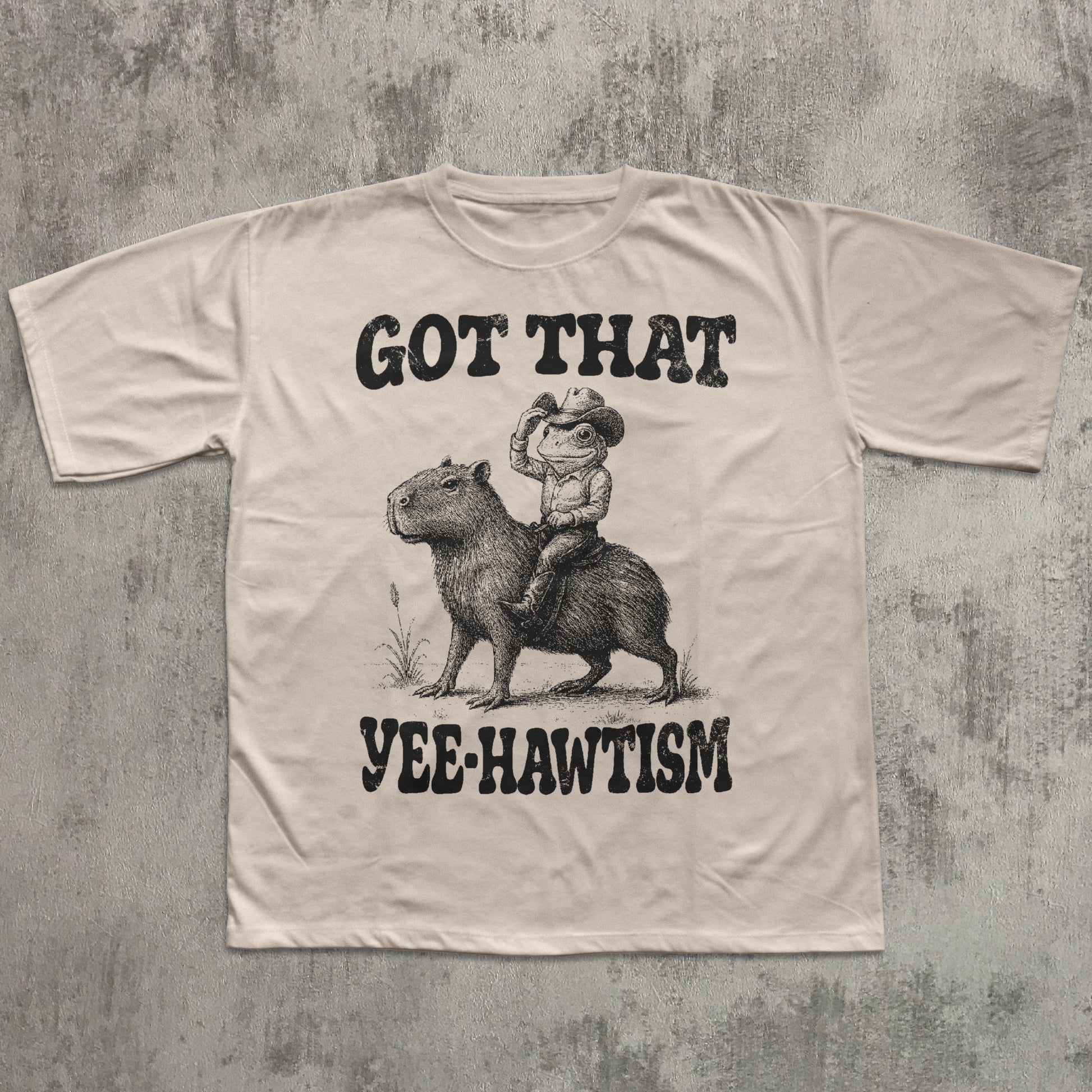 Beige t-shirt with graphic of a cowboy riding a capybara and text 'Got That Yee-Hawtism' on a textured gray background