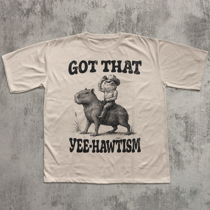Beige t-shirt with graphic of a cowboy riding a capybara and text 'Got That Yee-Hawtism' on a textured gray background