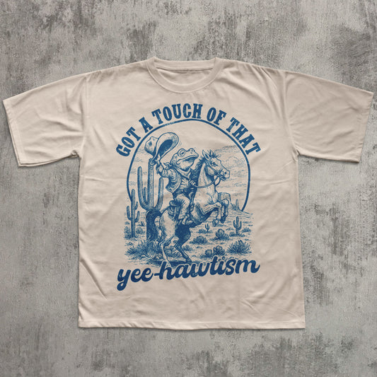 Natural T-shirt featuring a blue vintage-style illustration of a cowboy frog riding a rearing horse in a desert scene with cacti, with the text ‘Got a touch of that yee-hawtism’ above and below the graphic.