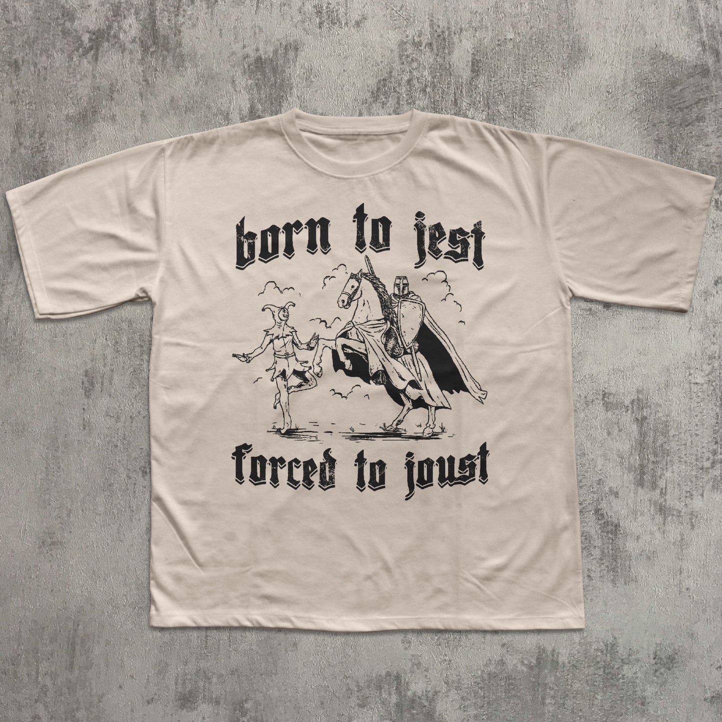 Beige t-shirt with black graphic and text 'born to jest, forced to joust' on a textured gray background