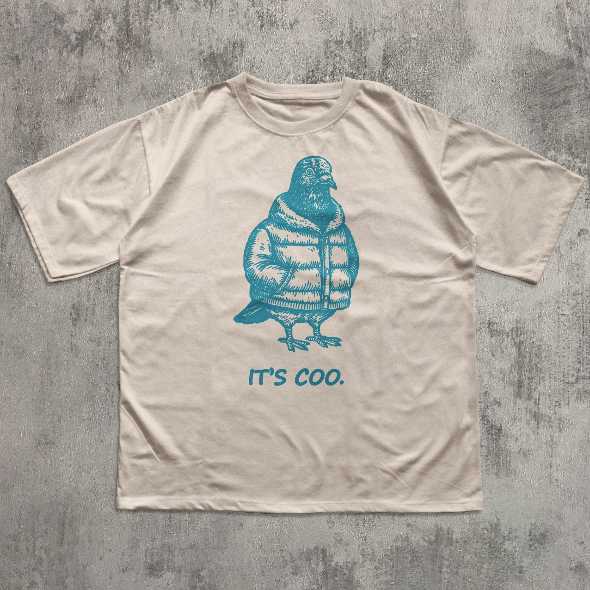 Beige t-shirt with blue penguin graphic and 'it's coo.' text on a textured gray background