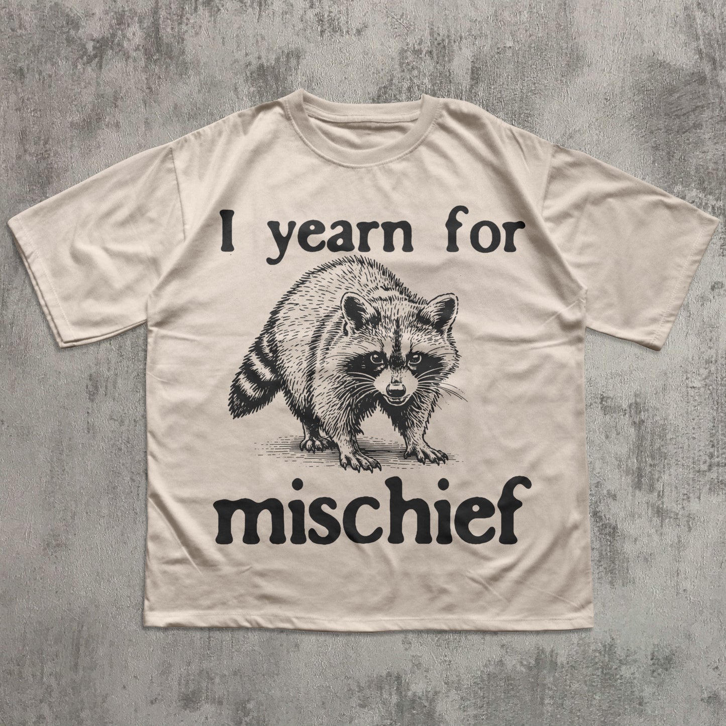 Beige t-shirt with a raccoon graphic and text 'I yearn for mischief' on a textured gray background