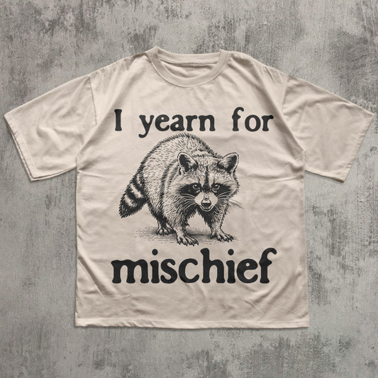 Beige t-shirt with a raccoon graphic and text 'I yearn for mischief' on a textured gray background