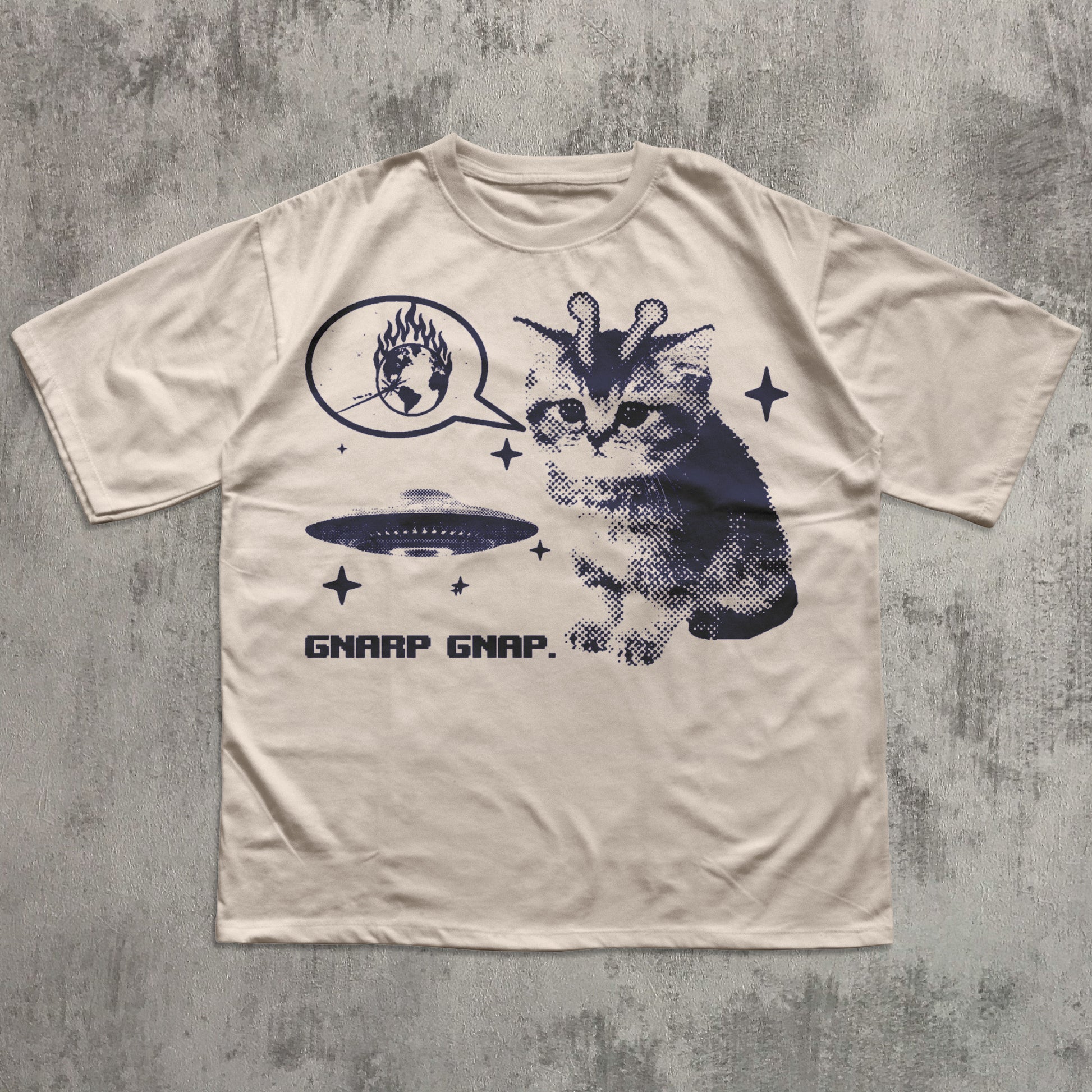 Beige t-shirt with a graphic of a cat and alien on a textured gray background