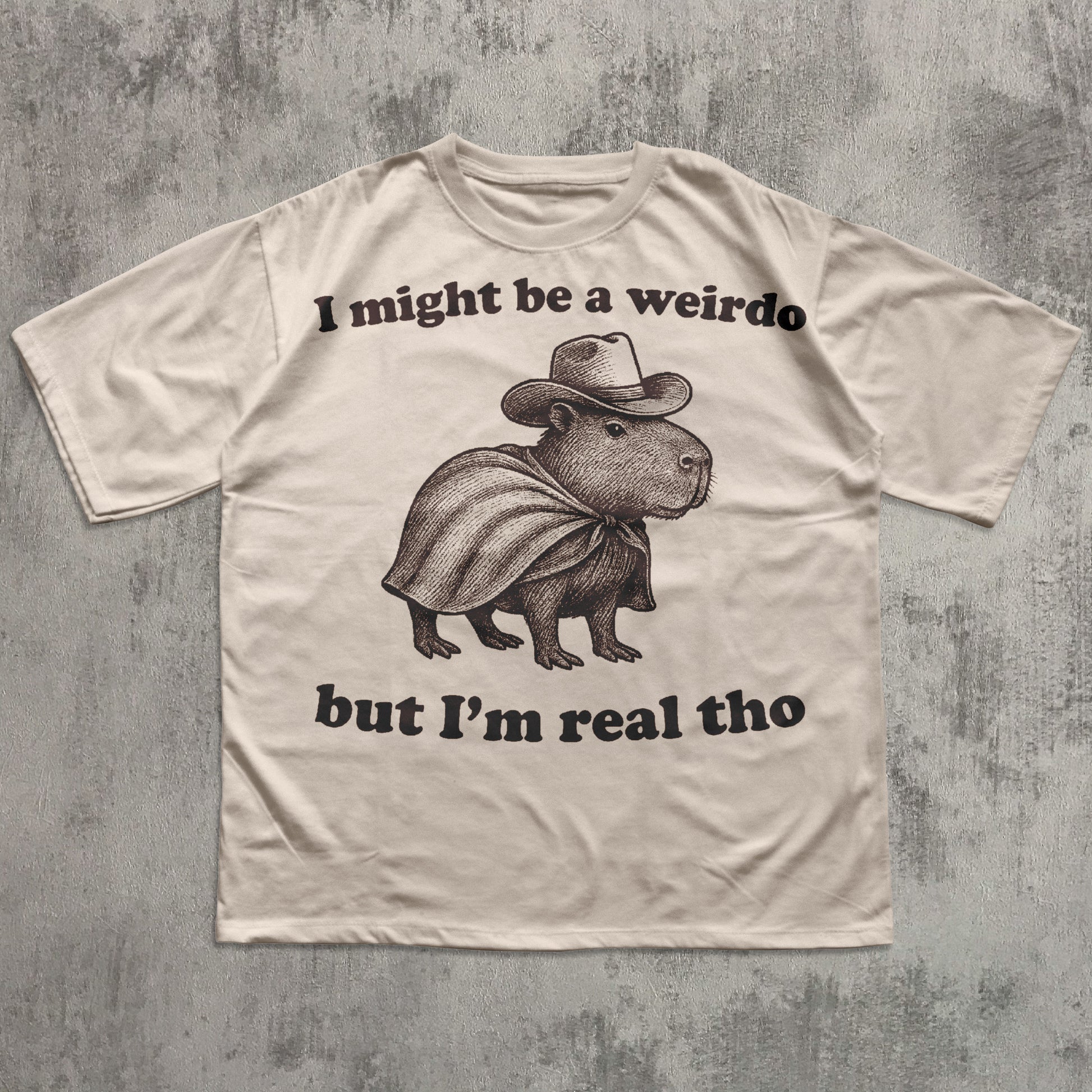 Beige t-shirt with a graphic of a capybara wearing a cowboy hat and cape, with text on a textured gray background.