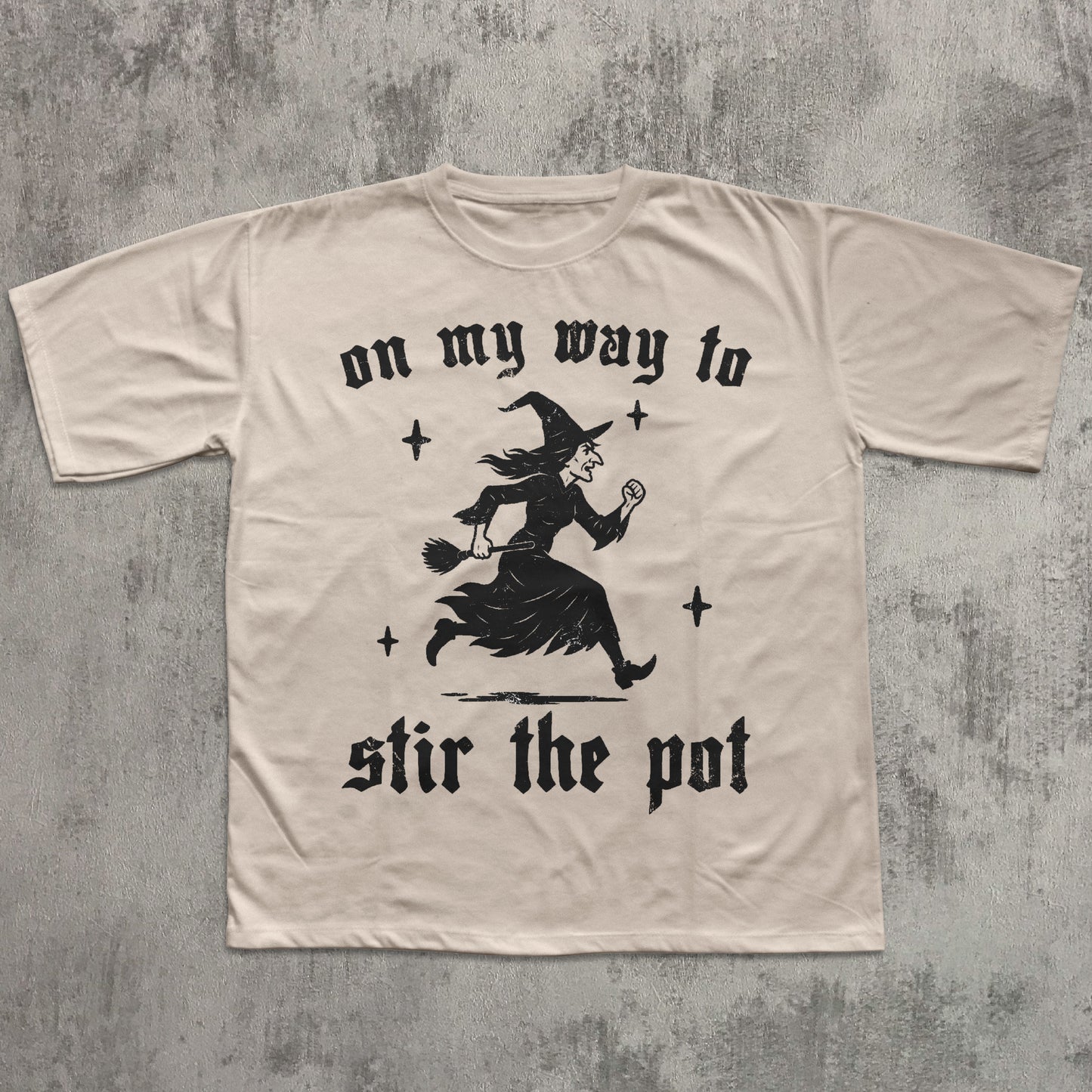 Beige t-shirt with black witch graphic and text on a textured gray background