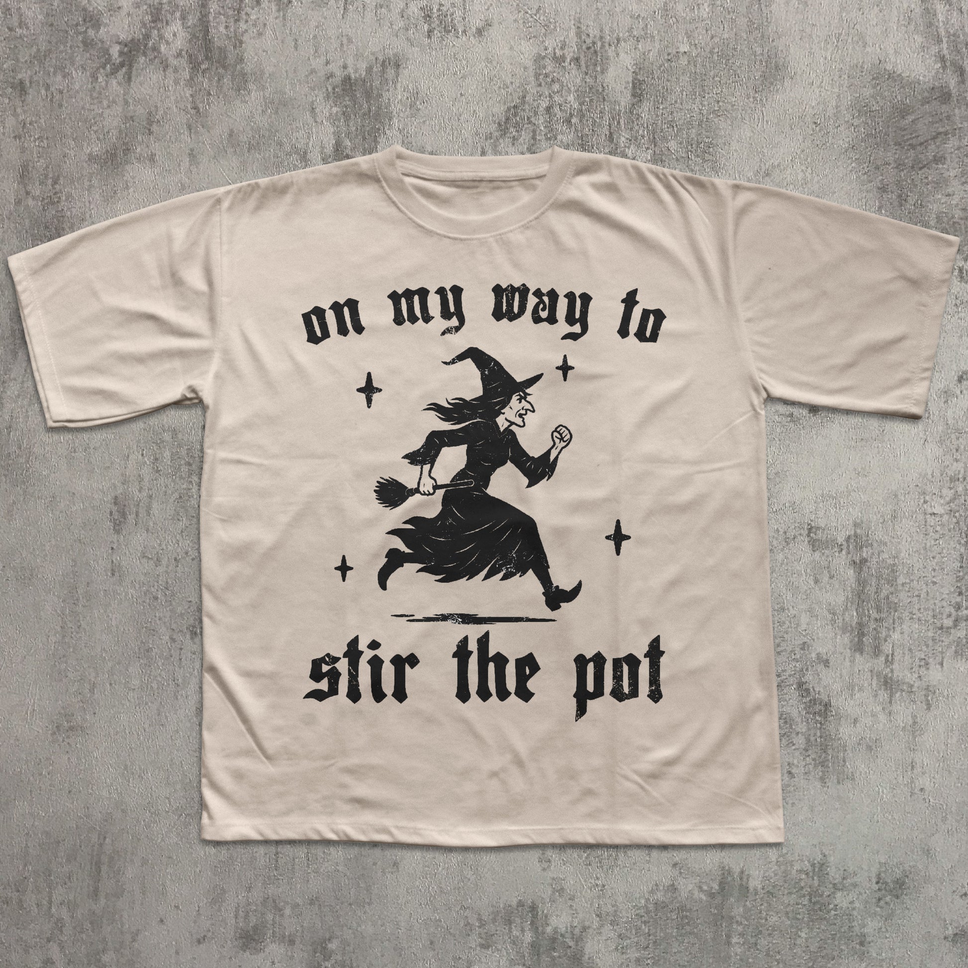 Beige t-shirt with black witch graphic and text on a textured gray background
