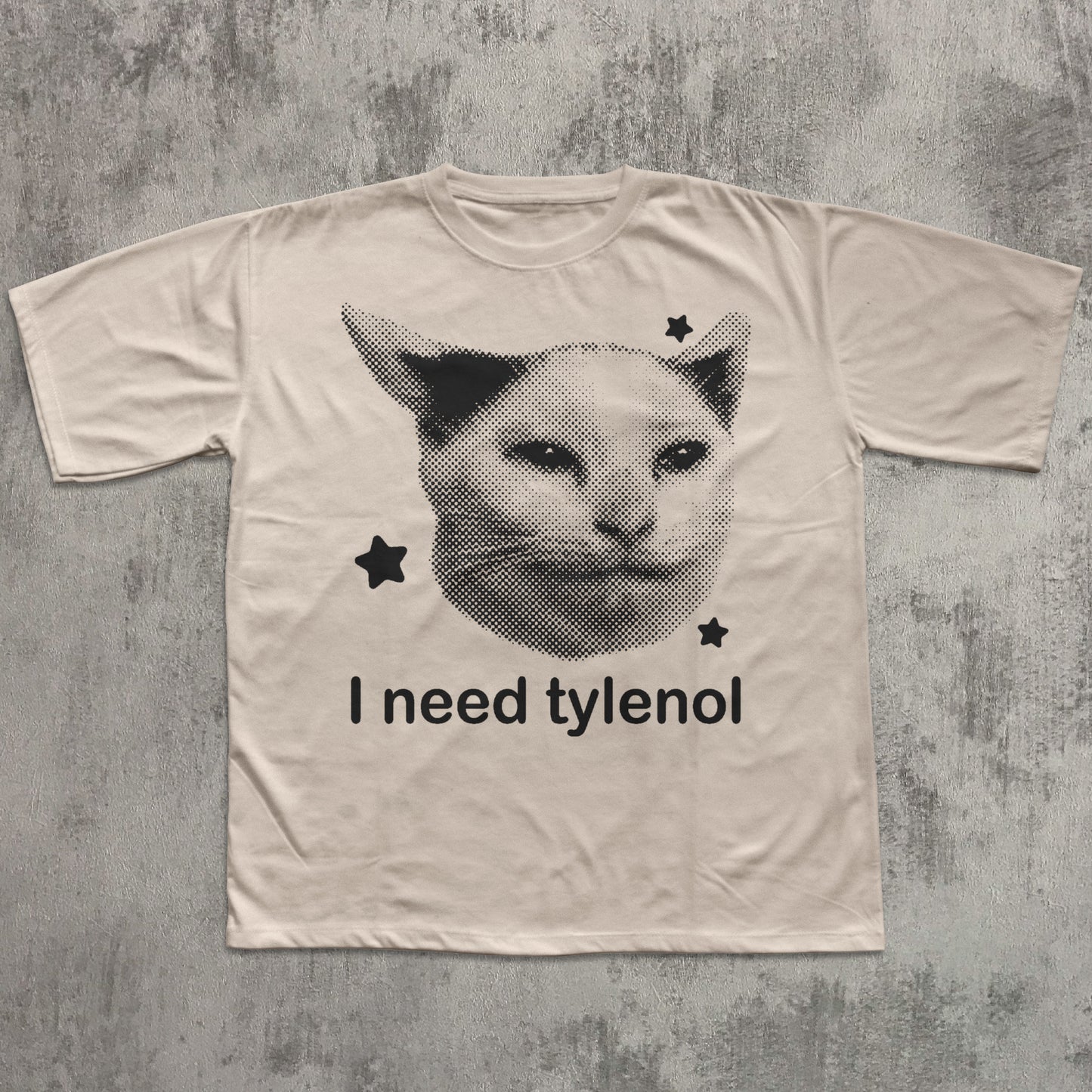 Beige t-shirt with a graphic of a cat's face and text 'I need tylenol' on a textured gray background