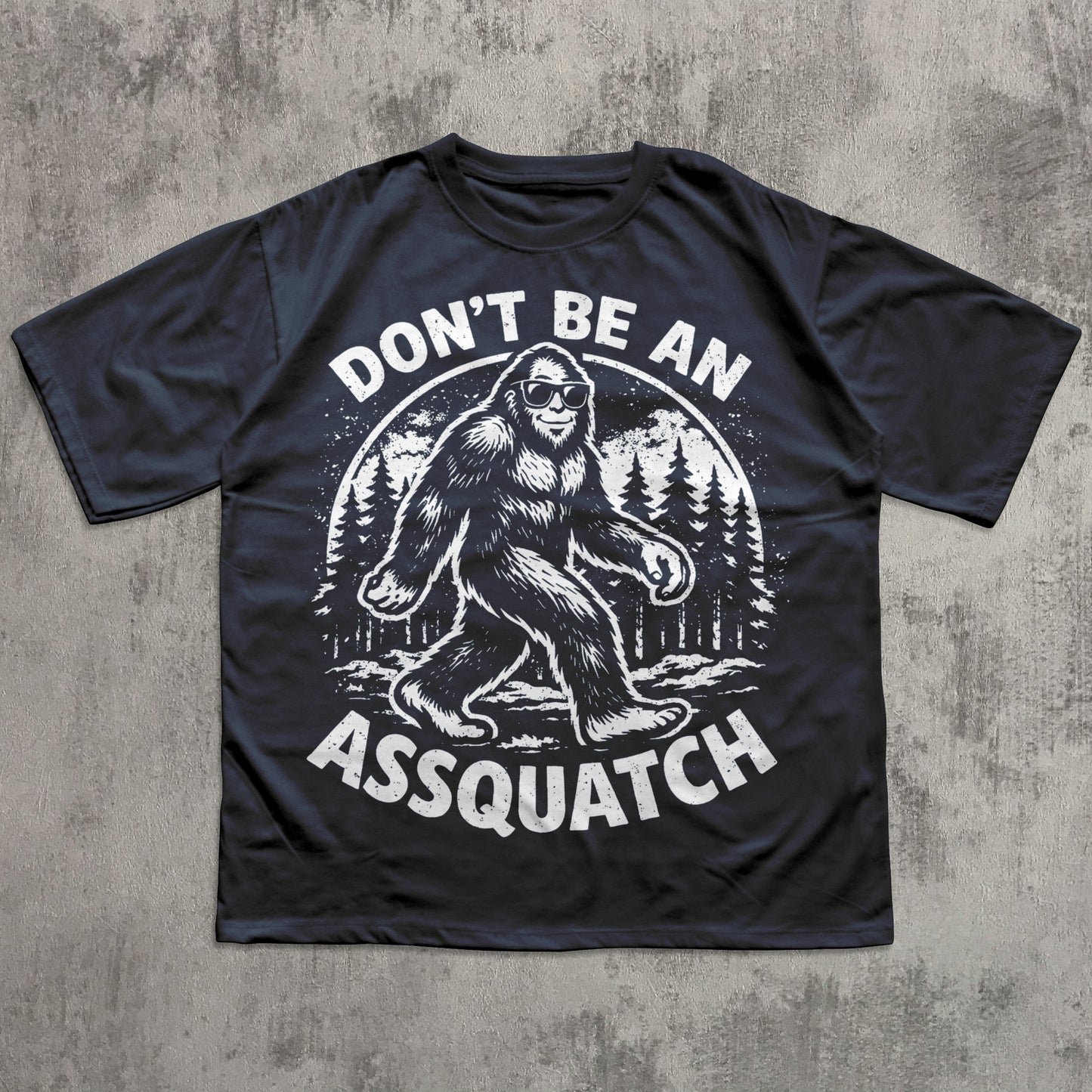 Black t-shirt with a graphic of a Sasquatch and text 'Don't Be An Assquatch' on a concrete surface.