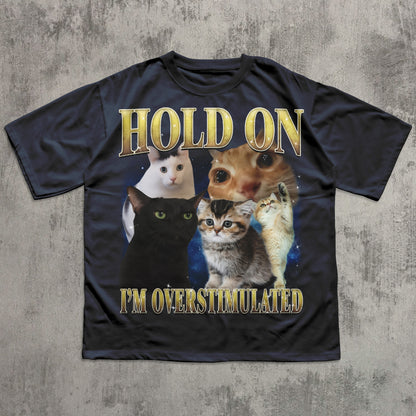 Black t-shirt with cartoon cats and text 'Hold on I'm overstimulated' on a textured gray background