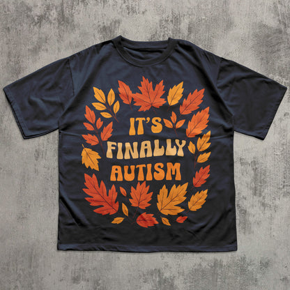 Black t-shirt with autumn leaf design and 'It's Finally Autism' text on a concrete surface
