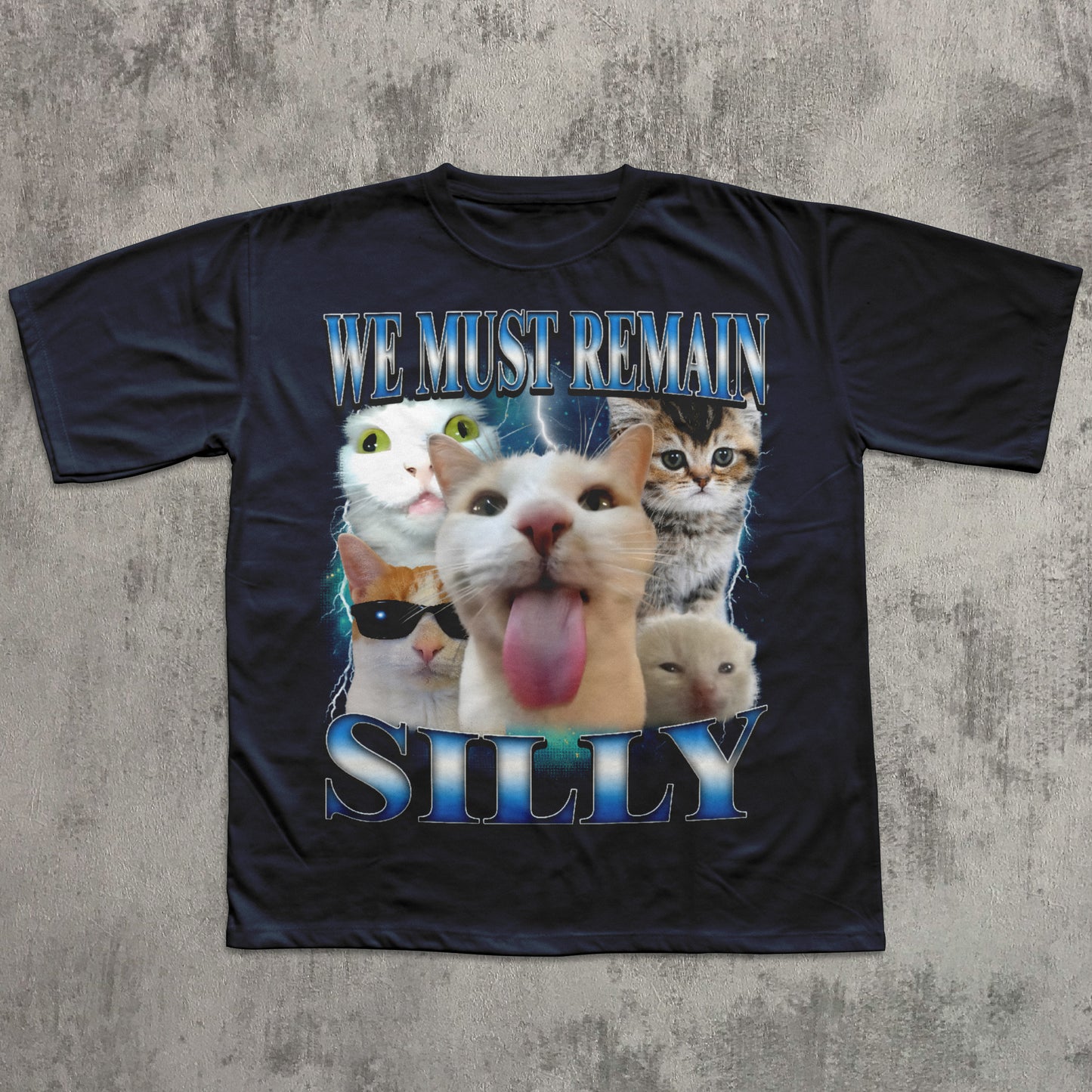 Black t-shirt with cartoon cats and text 'WE MUST REMAIN SILLY' on a textured gray background