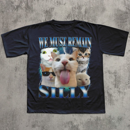 Black t-shirt with cartoon cats and text 'WE MUST REMAIN SILLY' on a textured gray background