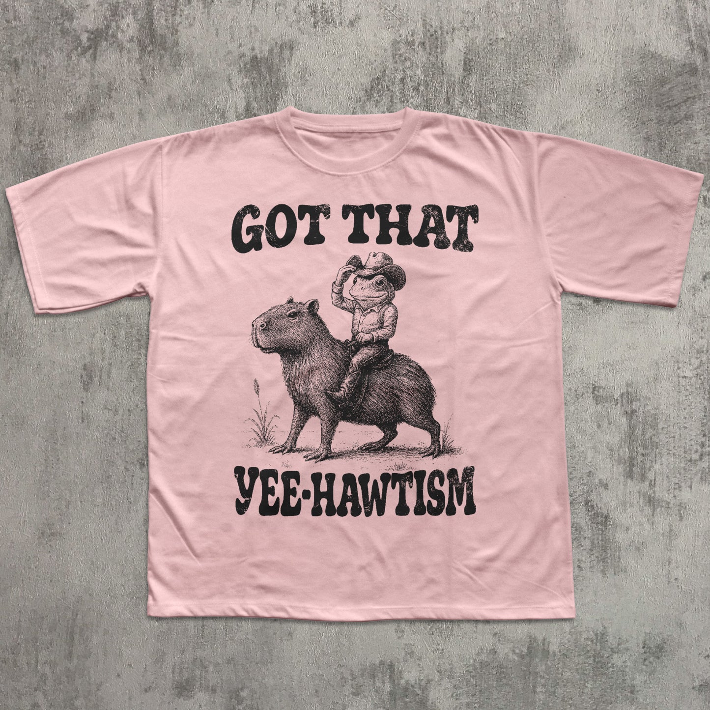 Pink t-shirt with a graphic of a cowboy riding a capybara and text on a concrete background