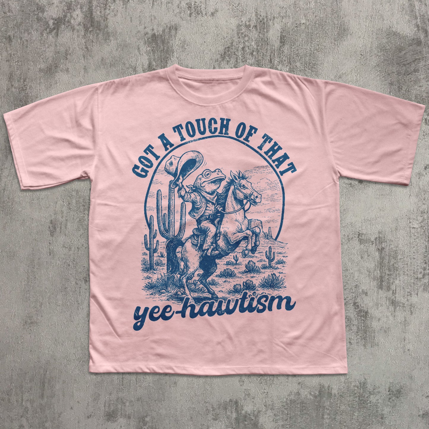 Pink T-shirt featuring a blue vintage-style illustration of a cowboy frog riding a rearing horse in a desert scene with cacti, with the text ‘Got a touch of that yee-hawtism’ above and below the graphic.