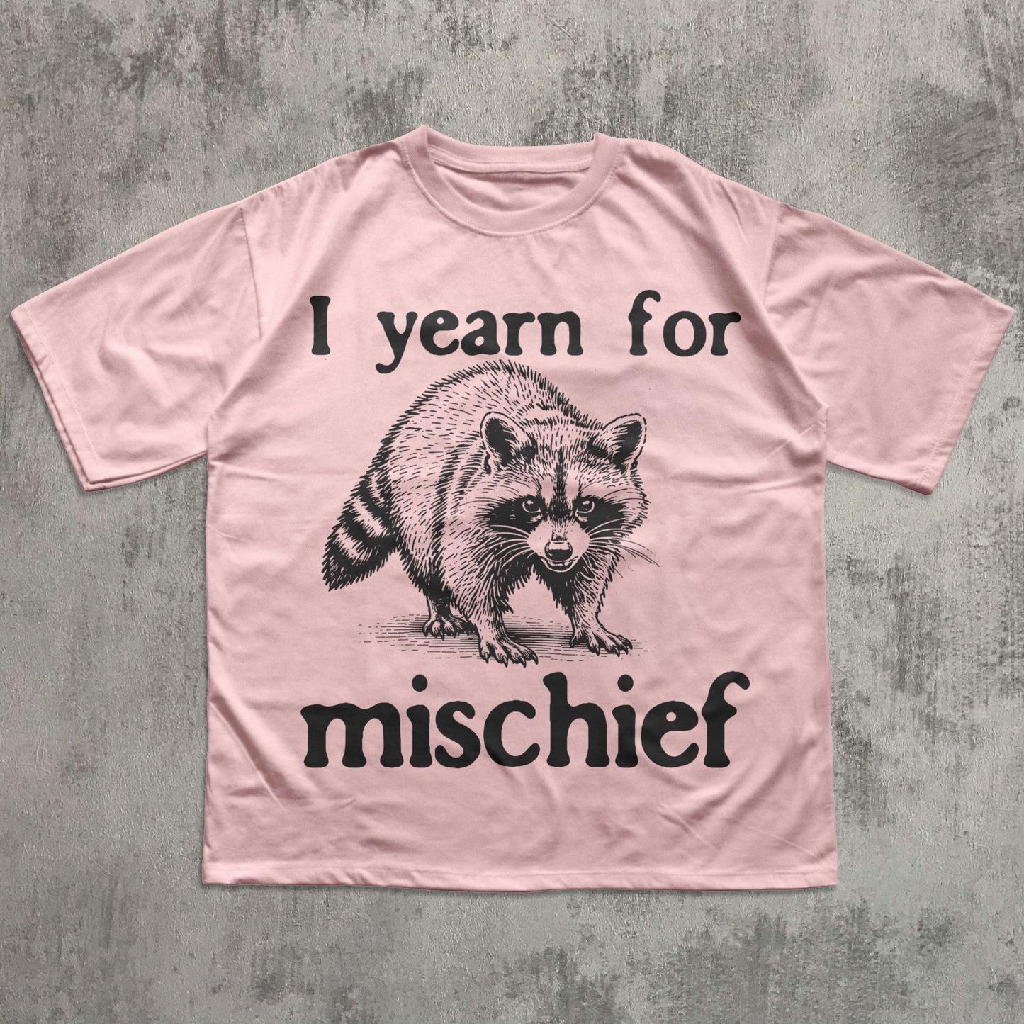 Pink t-shirt with raccoon graphic and text 'I yearn for mischief' on a textured gray background