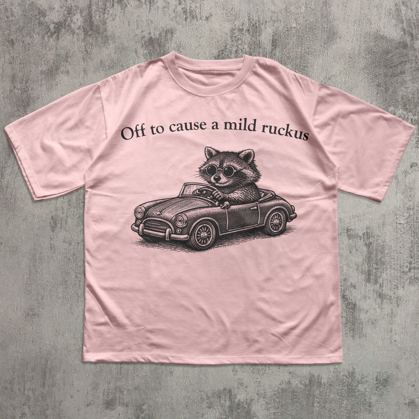 Pink t-shirt with a raccoon driving a car graphic and text on a textured gray background