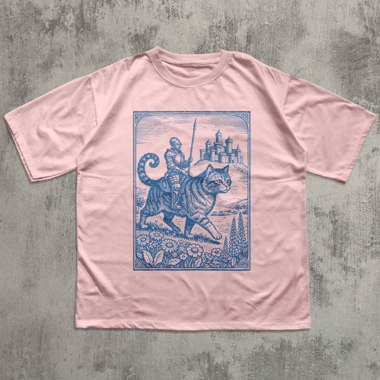 Pink t-shirt with blue graphic of a warrior on a cat with a castle in the background, on a textured gray surface.