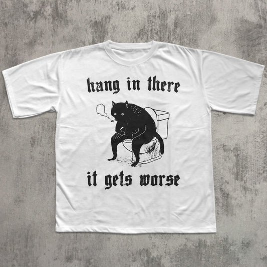 White t-shirt with black cat graphic and text 'hang in there it gets worse' on a textured gray background