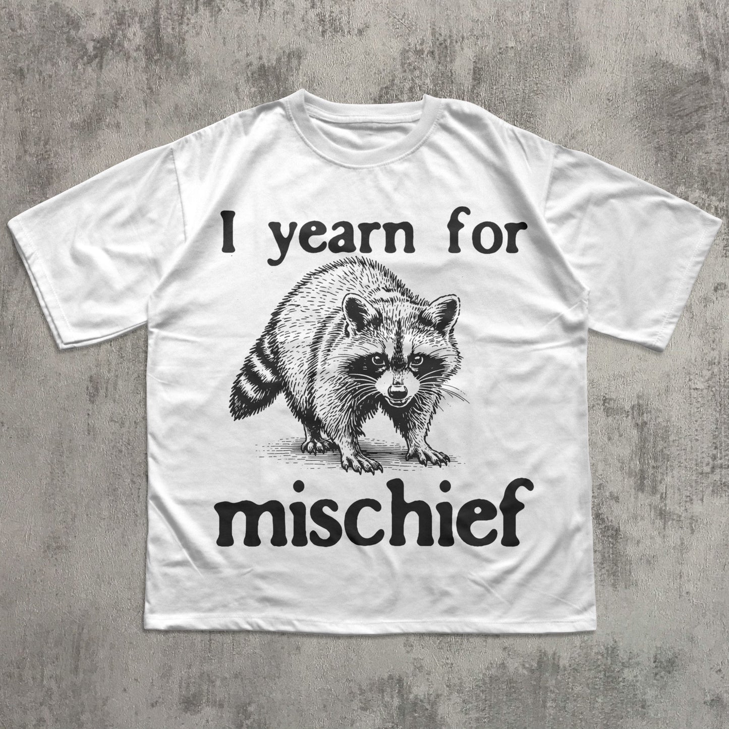 White t-shirt with a raccoon graphic and text 'I yearn for mischief' on a concrete surface.