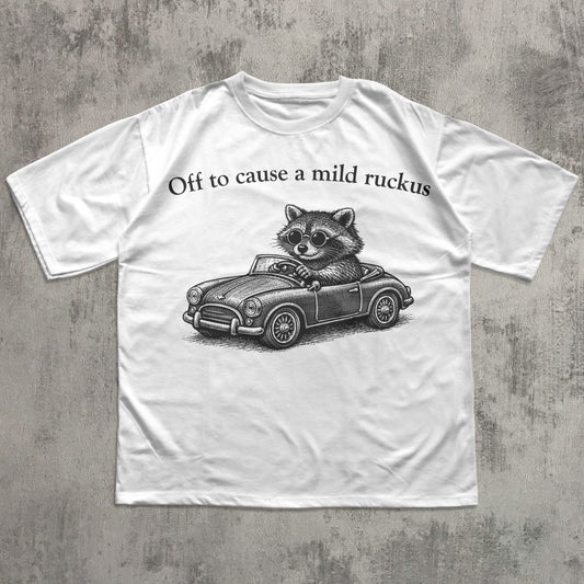 White t-shirt with a raccoon driving a car graphic and text on a concrete surface