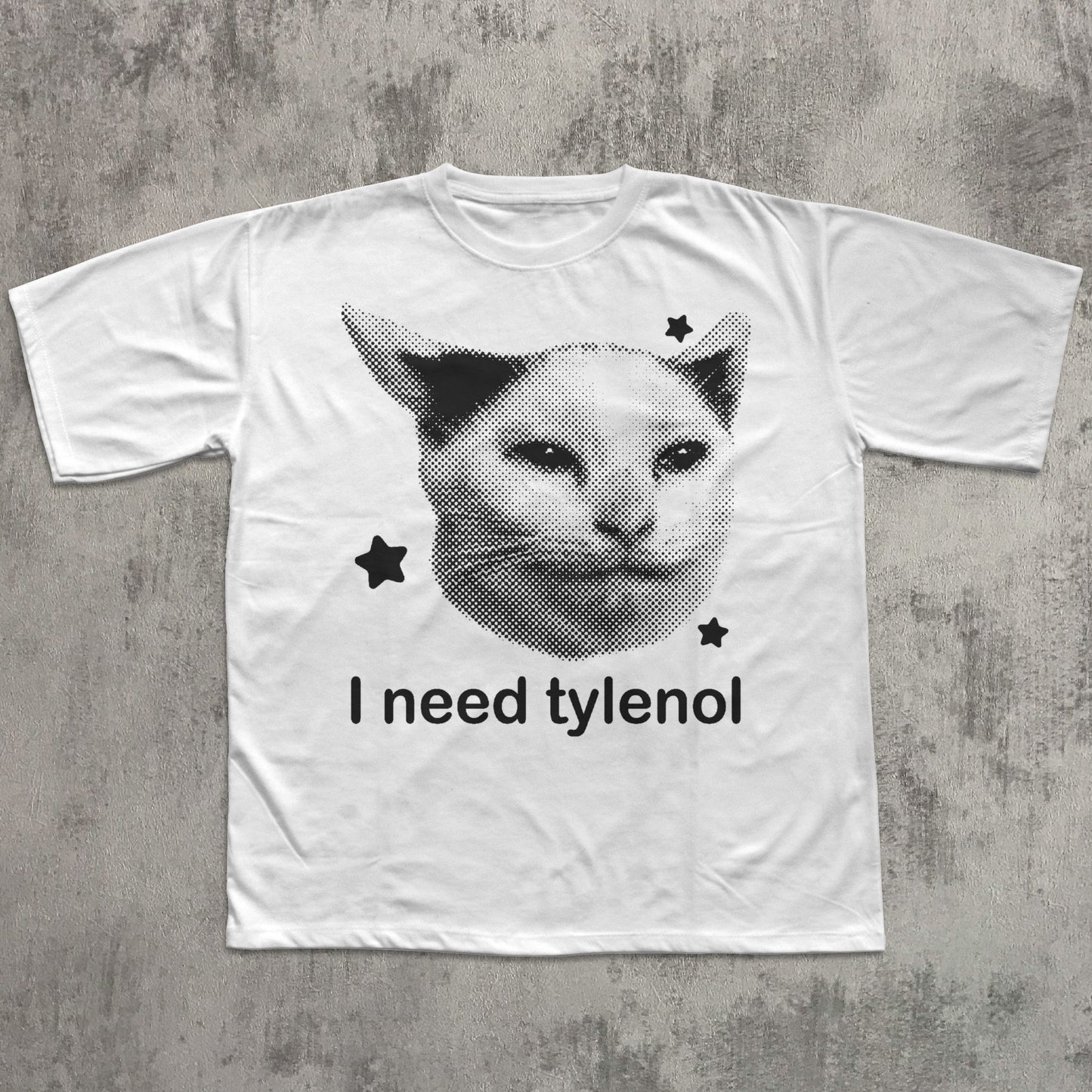 White t-shirt with a graphic of a cat's face and text 'I need tylenol' on a textured gray background