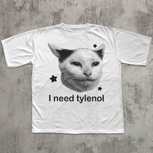 White t-shirt with a graphic of a cat's face and text 'I need tylenol' on a textured gray background